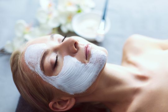 Mujer con una máscara facial blanca, ojos cerrados, relajándose en una mesa; entorno de spa.