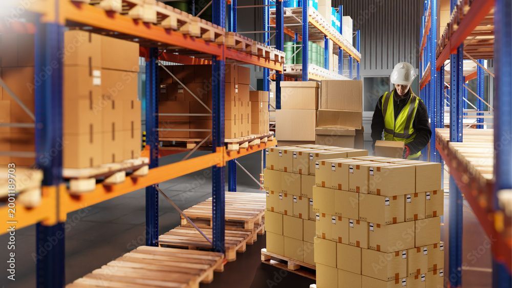Un ouvrier d'entrepôt portant un casque de chantier empile des cartons à côté de hautes étagères industrielles.