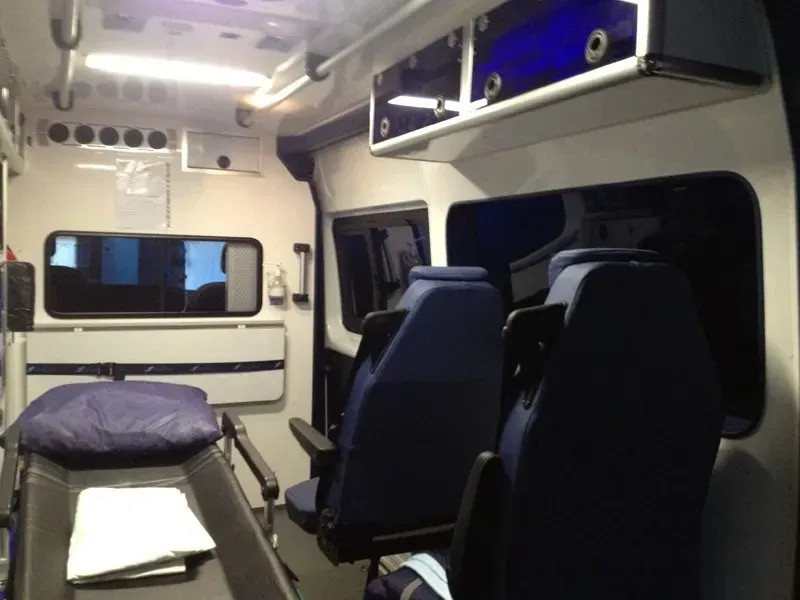 Intérieur d'une ambulance. On distingue deux sièges bleus, des armoires, une civière et des fenêtres.