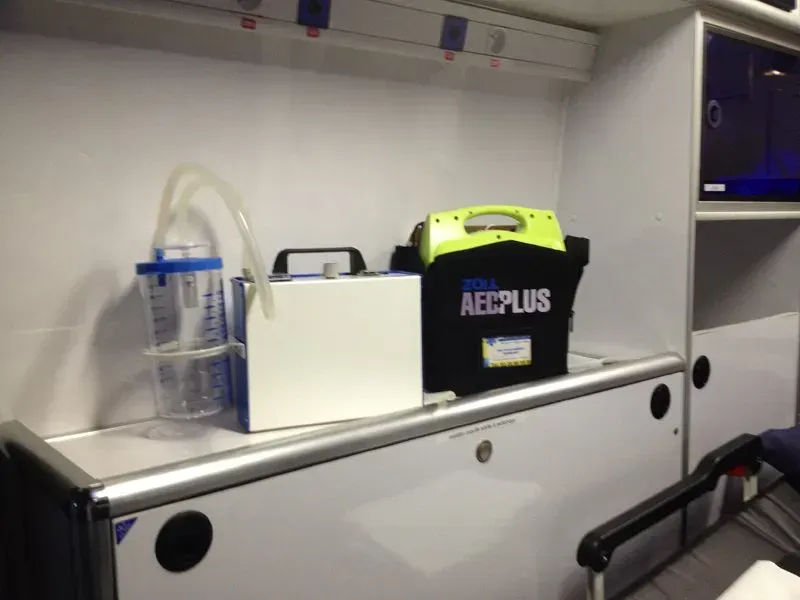À l'intérieur d'une ambulance, du matériel médical est posé sur une étagère. On aperçoit une bouteille d'oxygène et un défibrillateur. Blanc et noir.