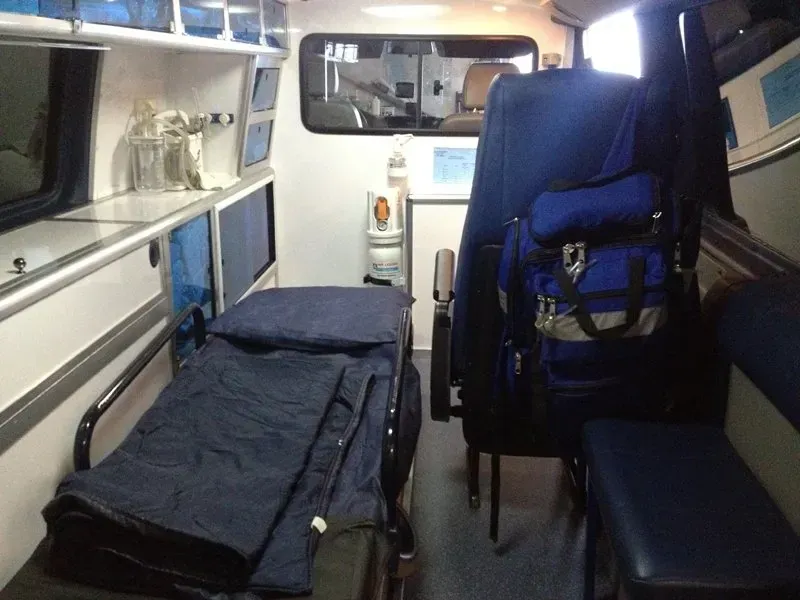 Intérieur d'une ambulance. Une civière est à gauche, un sac bleu est accroché à un siège à droite. Armoires et matériel.