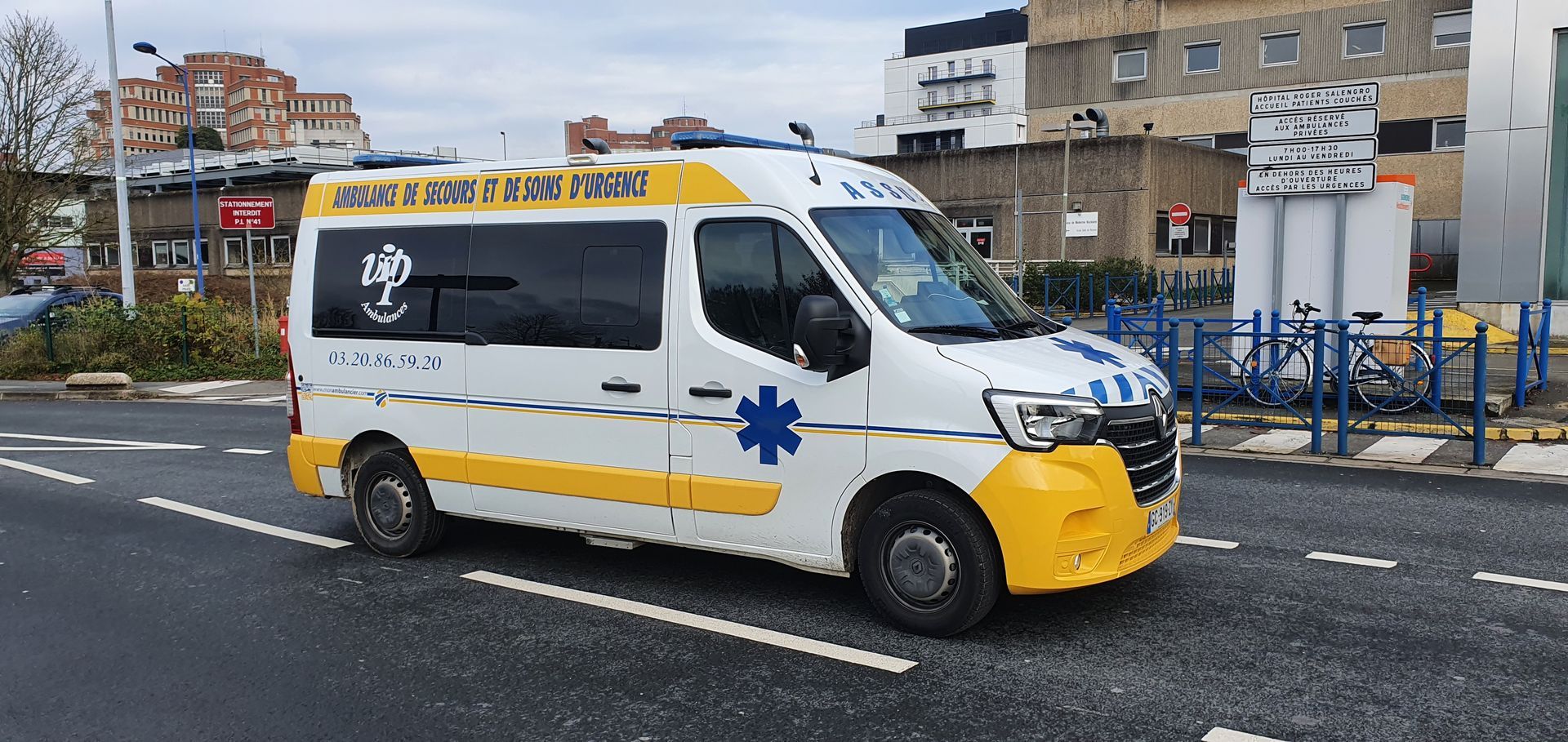 Une ambulance blanche avec des touches de jaune et un emblème en forme d'étoile bleue, sur une route devant un bâtiment.