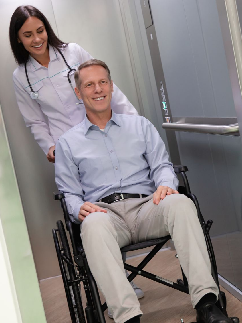 Un médecin pousse une personne en fauteuil roulant dans un ascenseur. Tous deux sourient.