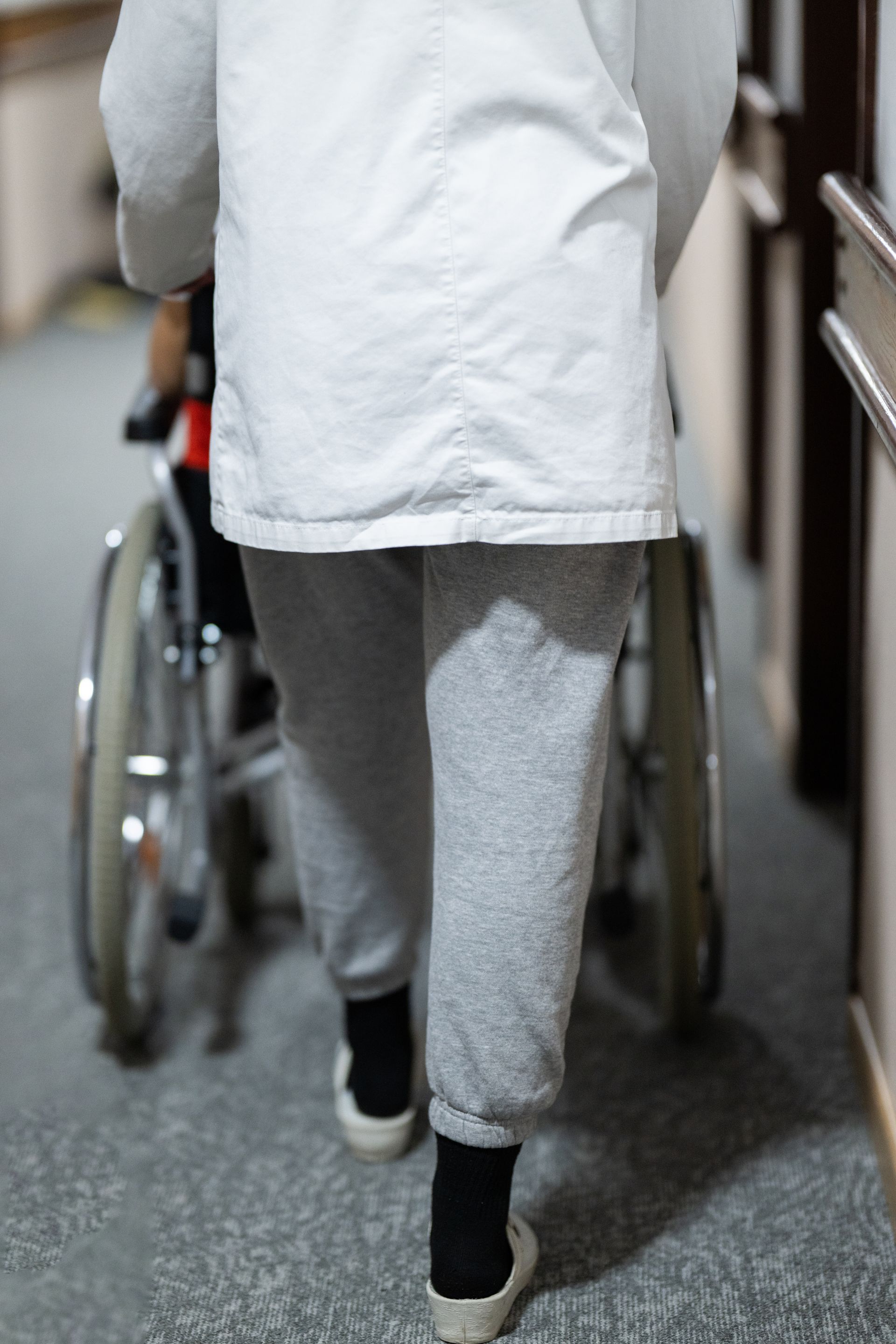 Une personne marche en poussant un fauteuil roulant dans un couloir. Elle porte un pantalon de survêtement gris, une veste blanche et des chaussures.