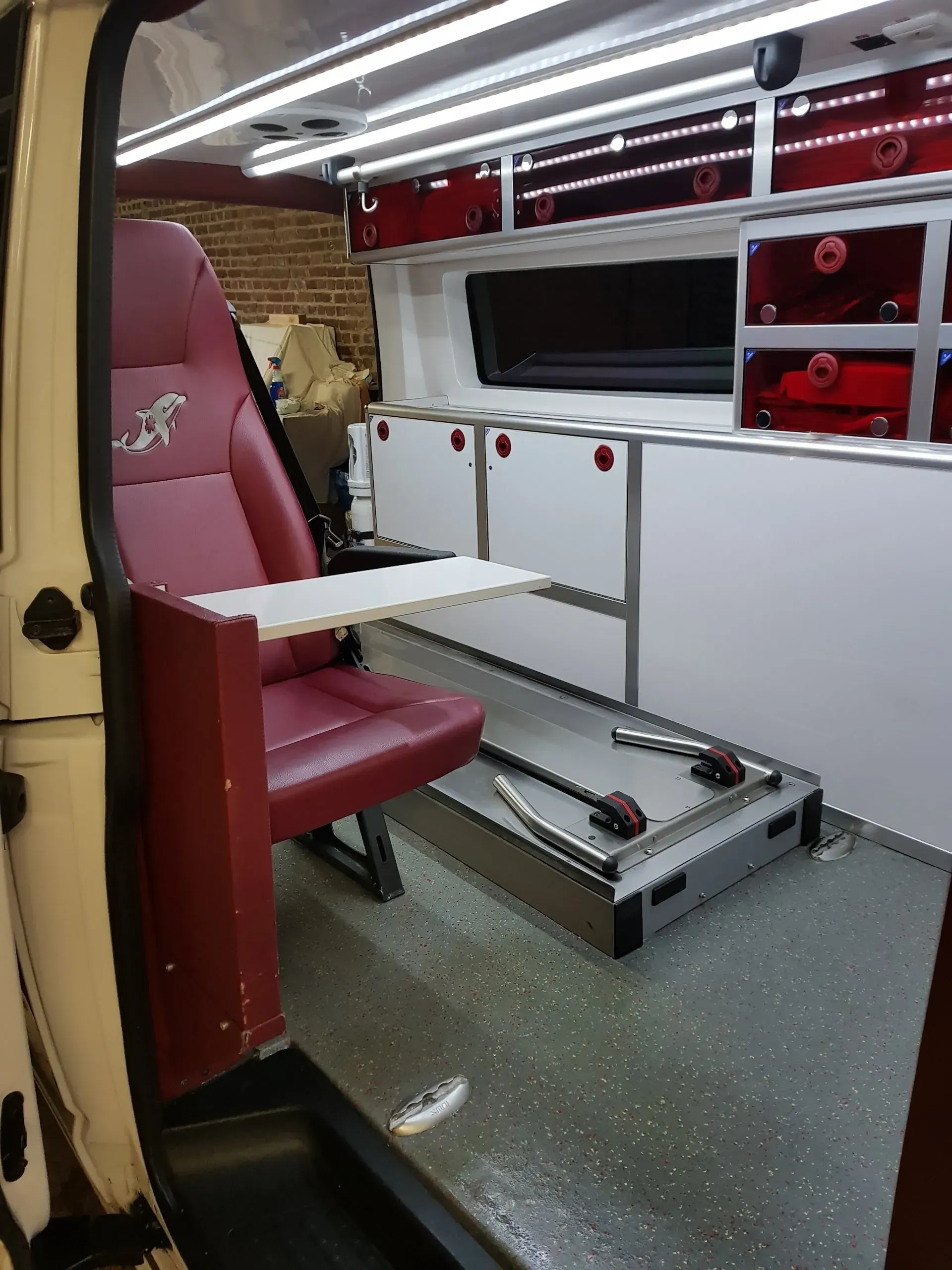 Intérieur d'une ambulance. Siège rouge avec tablette, placards blancs et compartiments de rangement rouges.