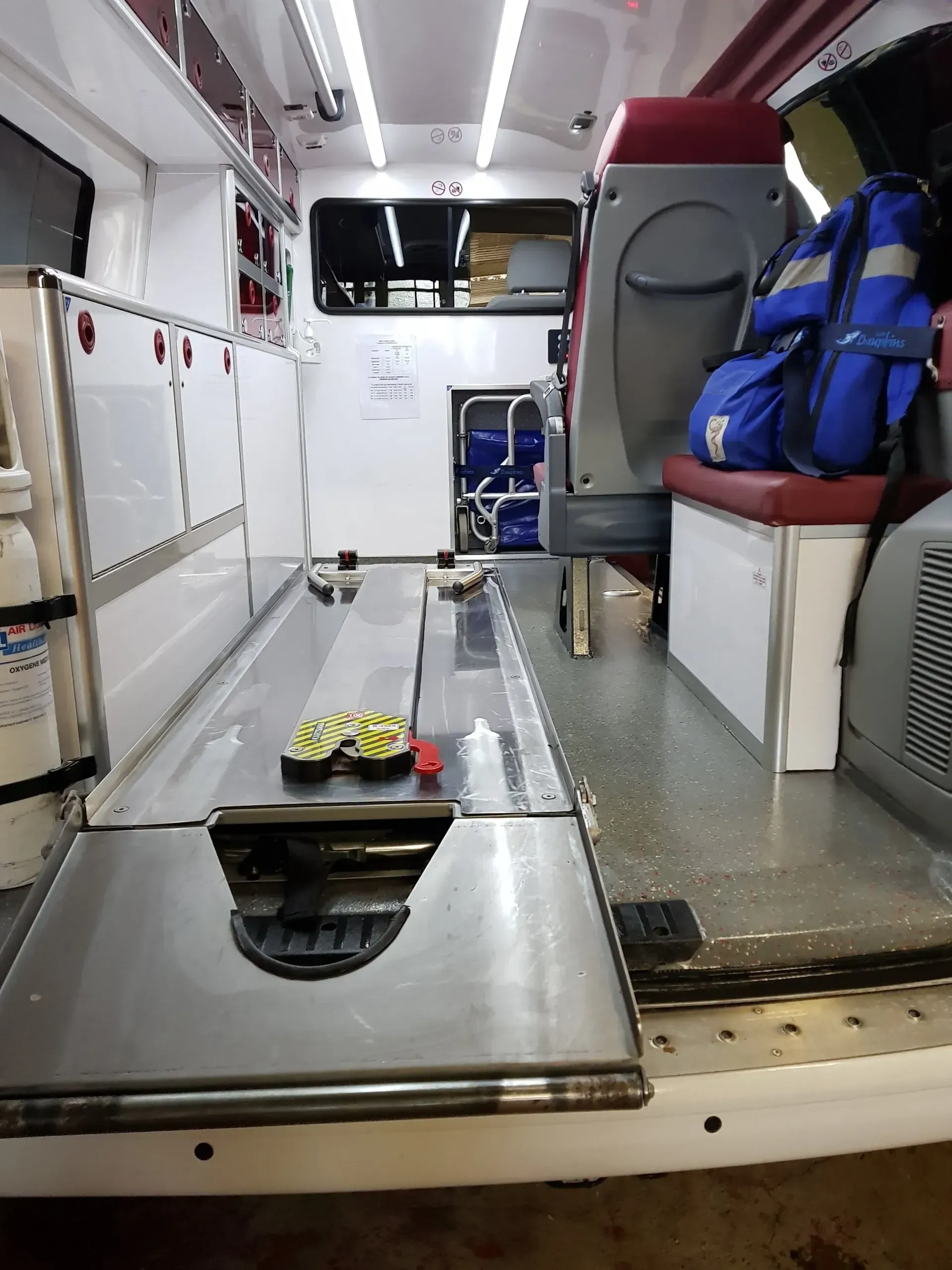 À l'intérieur d'une ambulance : sol en métal brillant, placards, banquette avec un sac à dos.