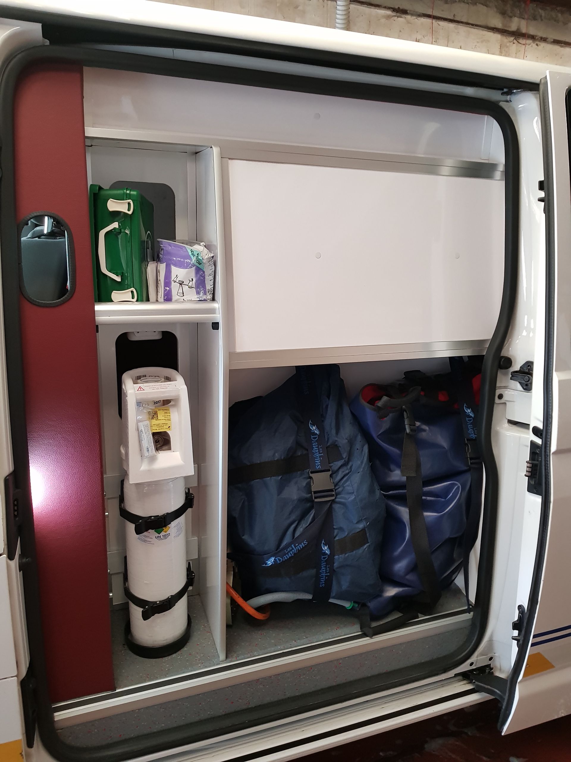 Intérieur d'un compartiment d'ambulance, montrant des étagères avec du matériel médical, une bouteille d'oxygène et des équipements de protection.