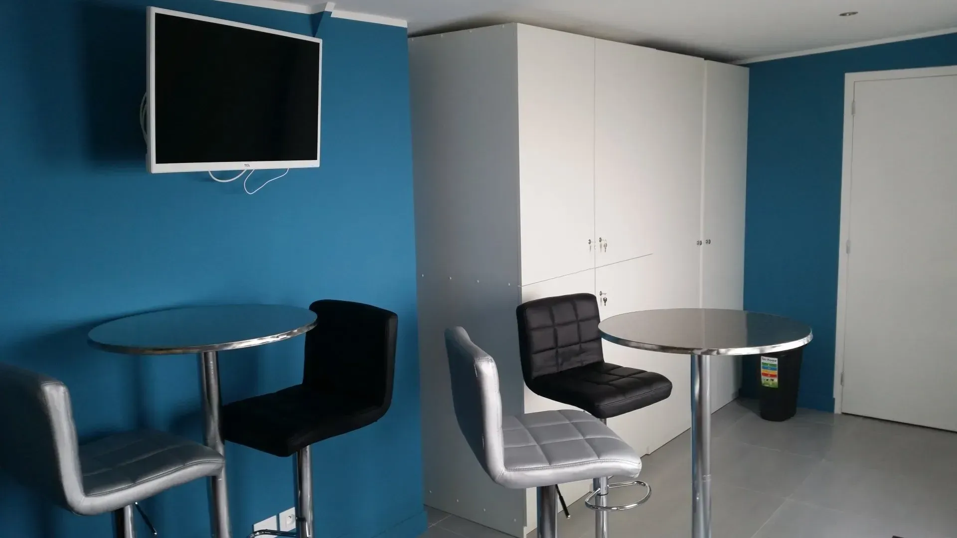 Deux tables rondes avec tabourets dans une pièce aux murs bleus et blancs ; un téléviseur est accroché au mur bleu.