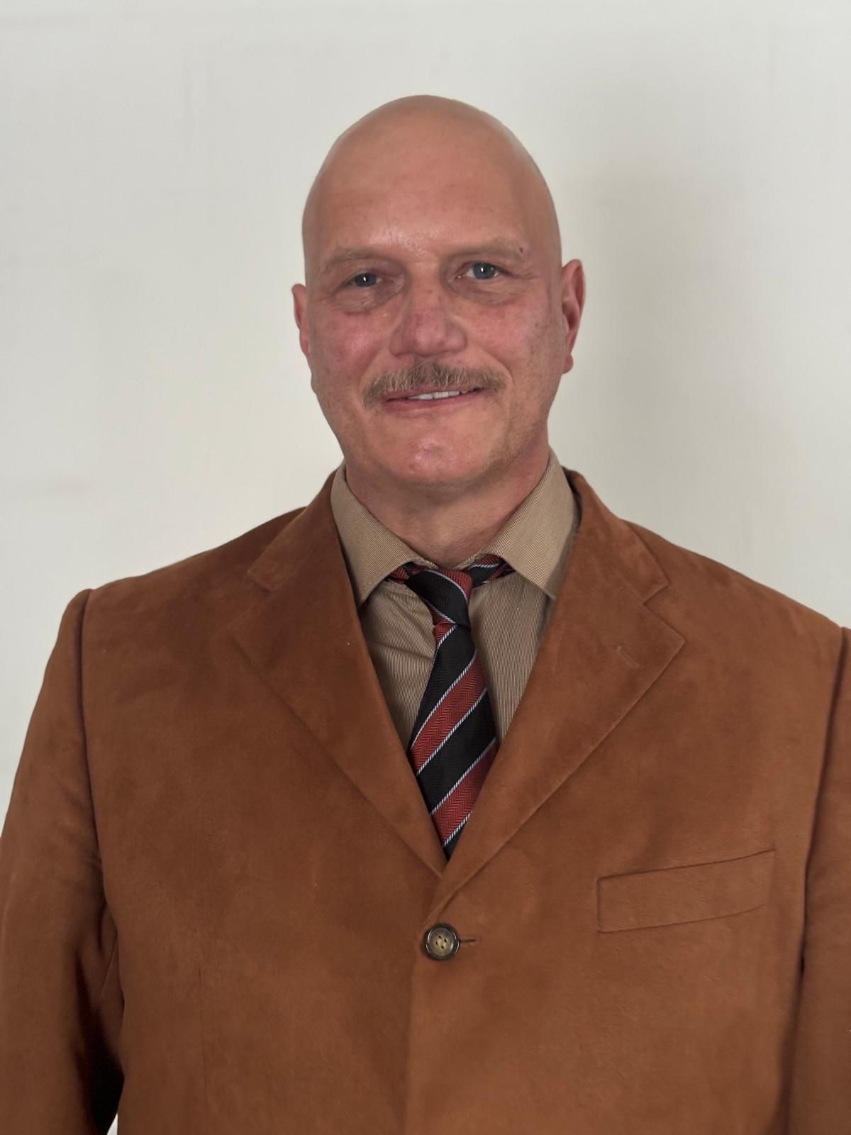 Ein kahlköpfiger Mann mit strengem Gesichtsausdruck, der ein bordeauxrotes Hemd und eine gestreifte Krawatte trägt.