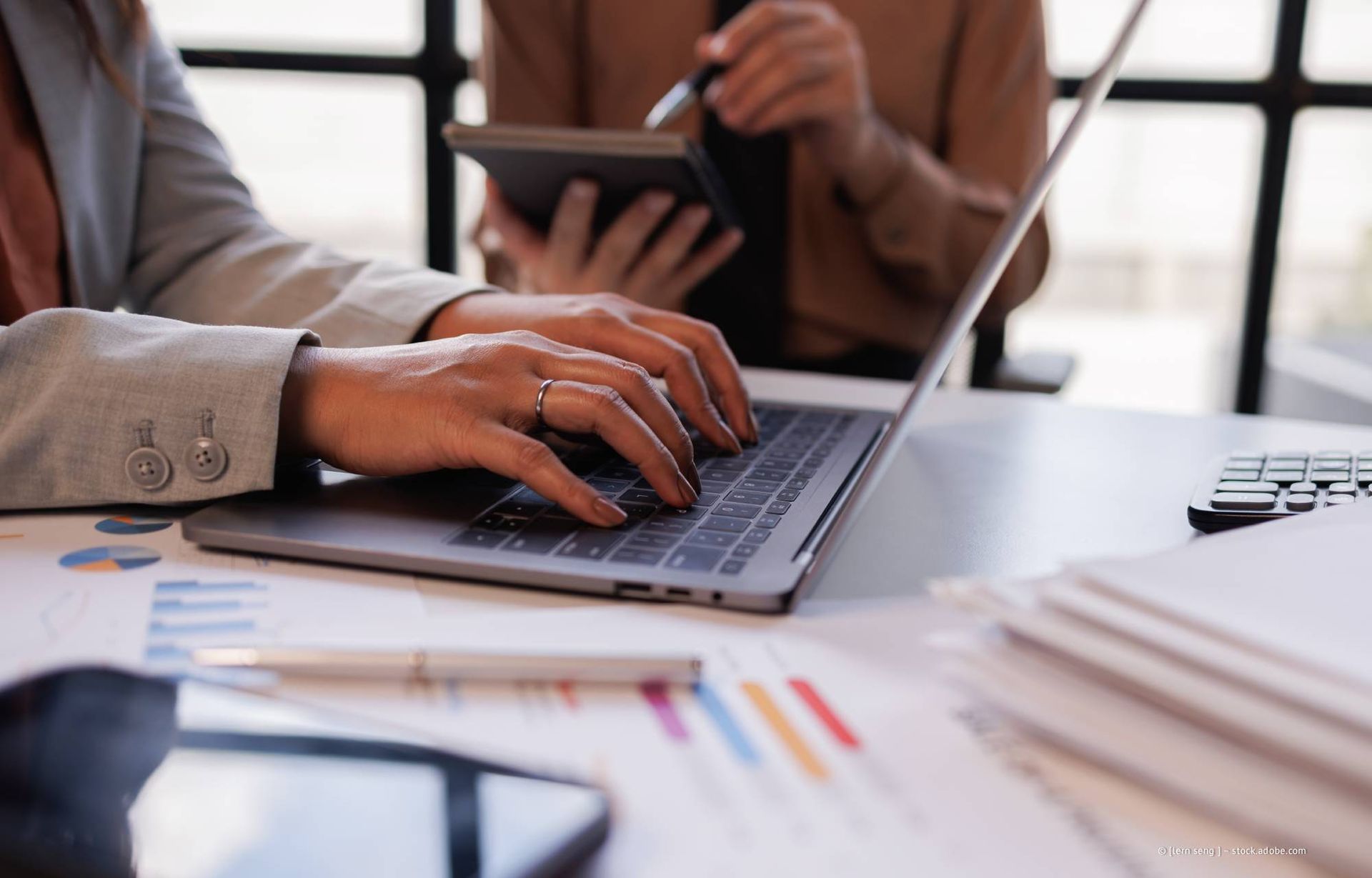 Hände tippen auf einem Laptop, im Hintergrund sind Diagramme und ein Tablet in einer Büroumgebung zu sehen.