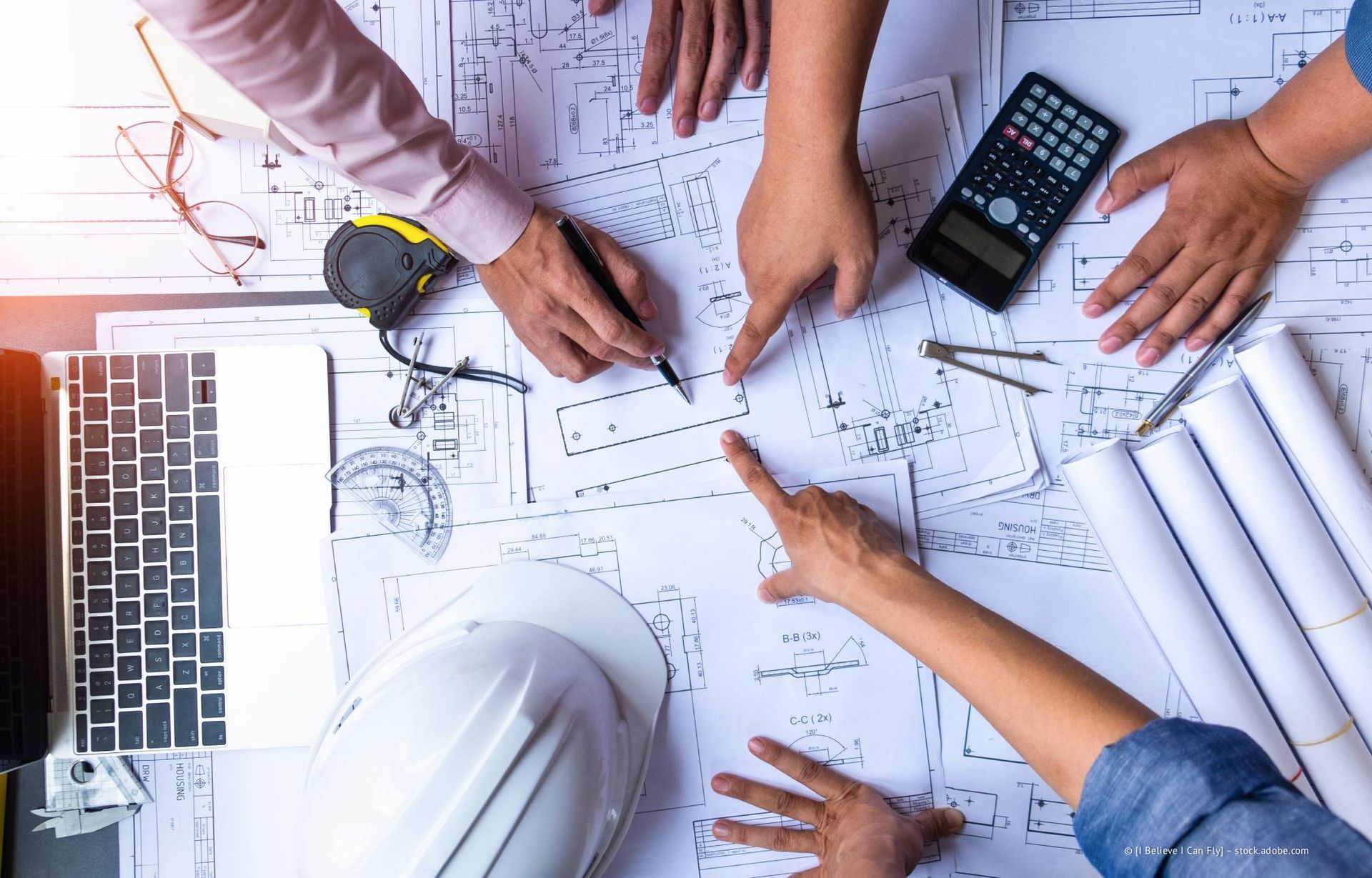 Hände zeigen auf Baupläne auf einem Schreibtisch, neben dem ein Schutzhelm, ein Taschenrechner und zusammengerollte Pläne liegen.