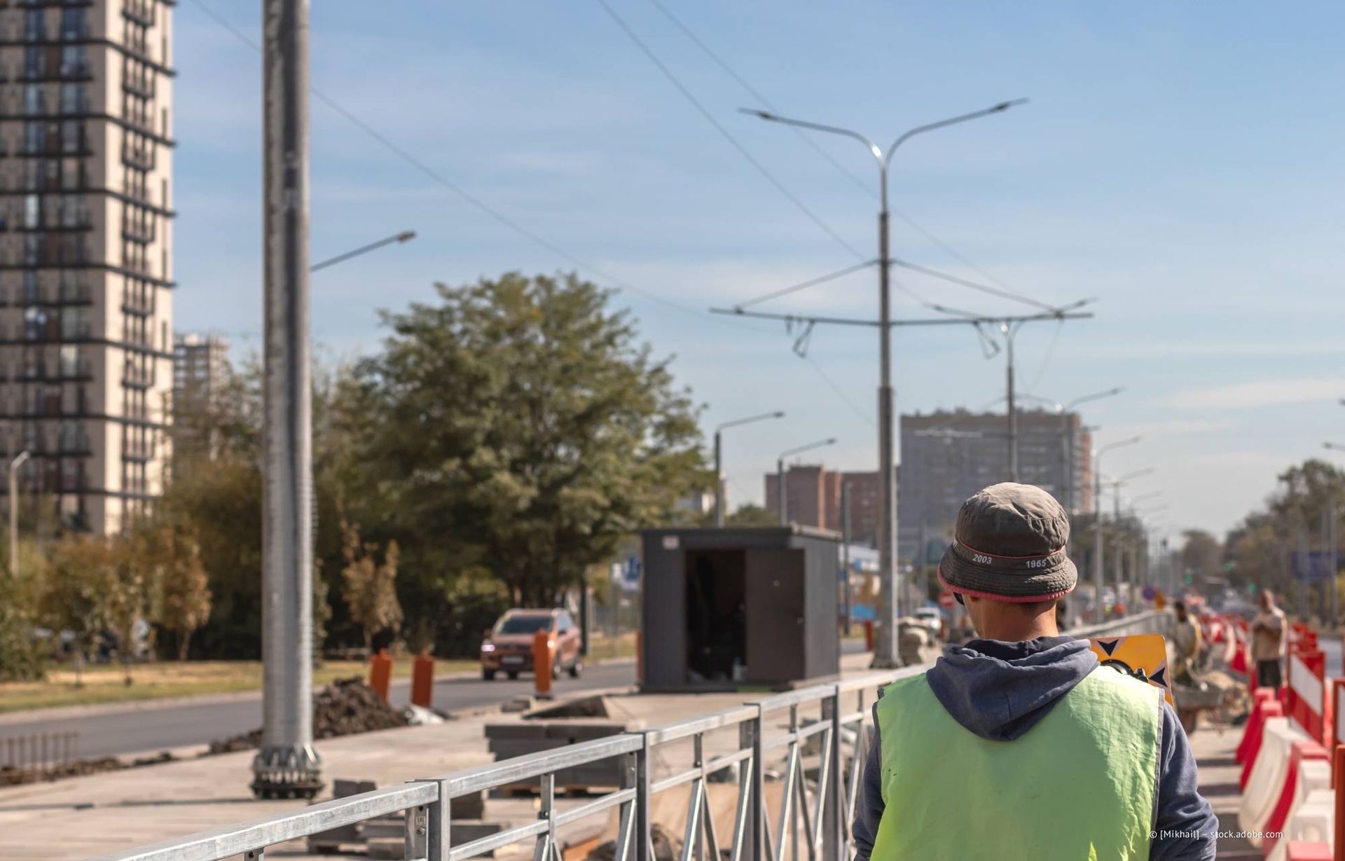Ein Bauarbeiter in Warnweste steht auf dem Bürgersteig neben einer Absperrung; im Hintergrund ist eine Straße mit Verkehr und Gebäuden zu sehen.
