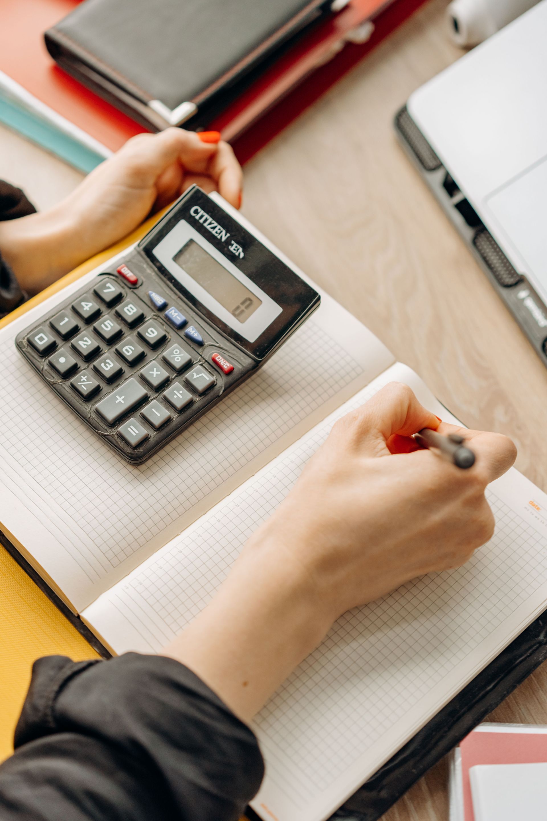 Manos escribiendo en un cuaderno junto a una calculadora sobre un escritorio con material de oficina