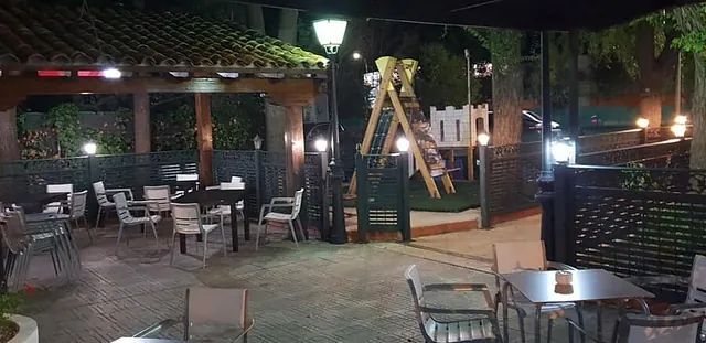 Comedor al aire libre en el patio por la noche con mesas, sillas, guirnaldas de luces y un parque infantil al fondo.