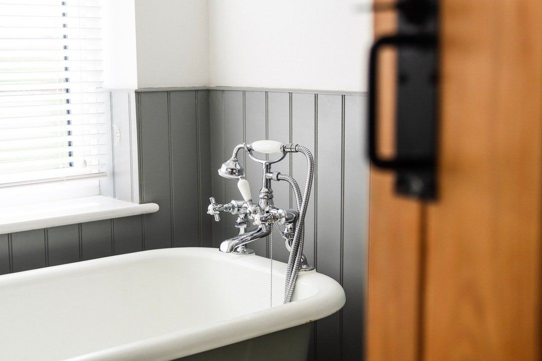 Bañera blanca con grifo cromado en un baño con paneles grises, junto a una ventana con persianas.