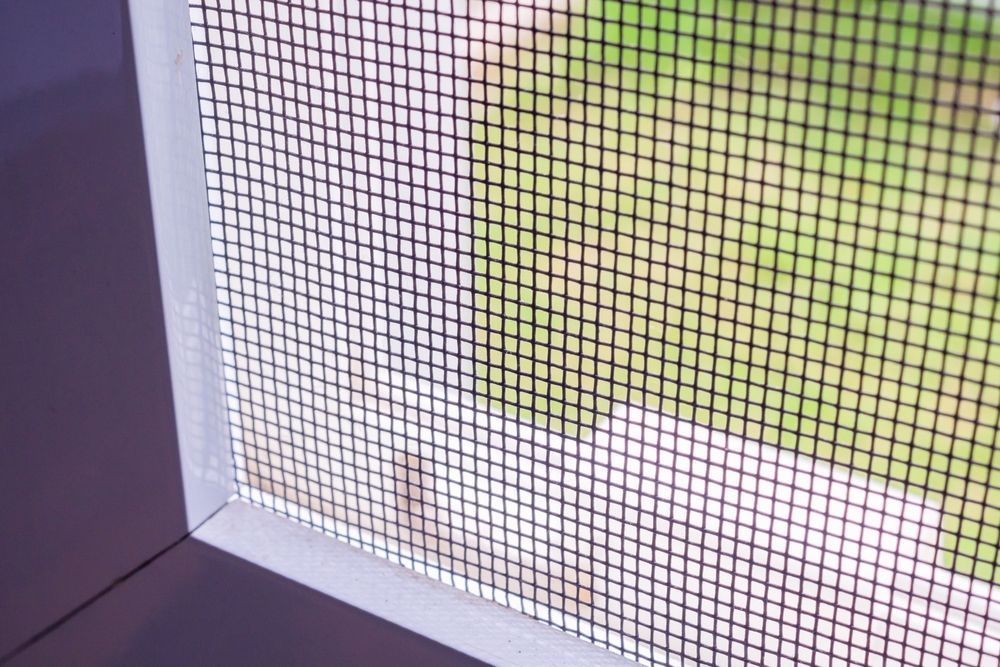 Primer plano de una mosquitera con follaje verde borroso en el exterior y un alféizar blanco.