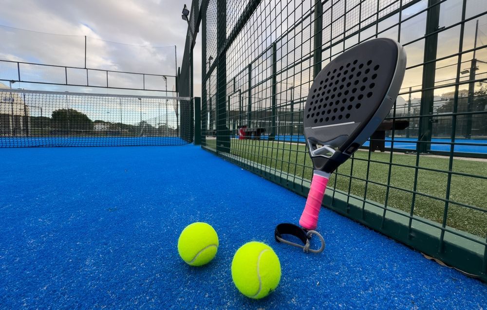 Una raqueta de pádel y dos pelotas amarillas sobre una pista azul junto a la red.