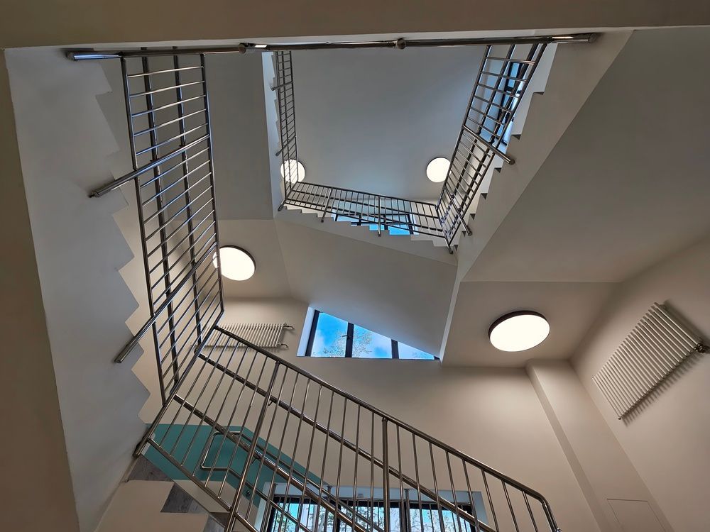 Vista de una escalera de varios pisos con barandillas metálicas, tragaluces y luces de techo redondas.