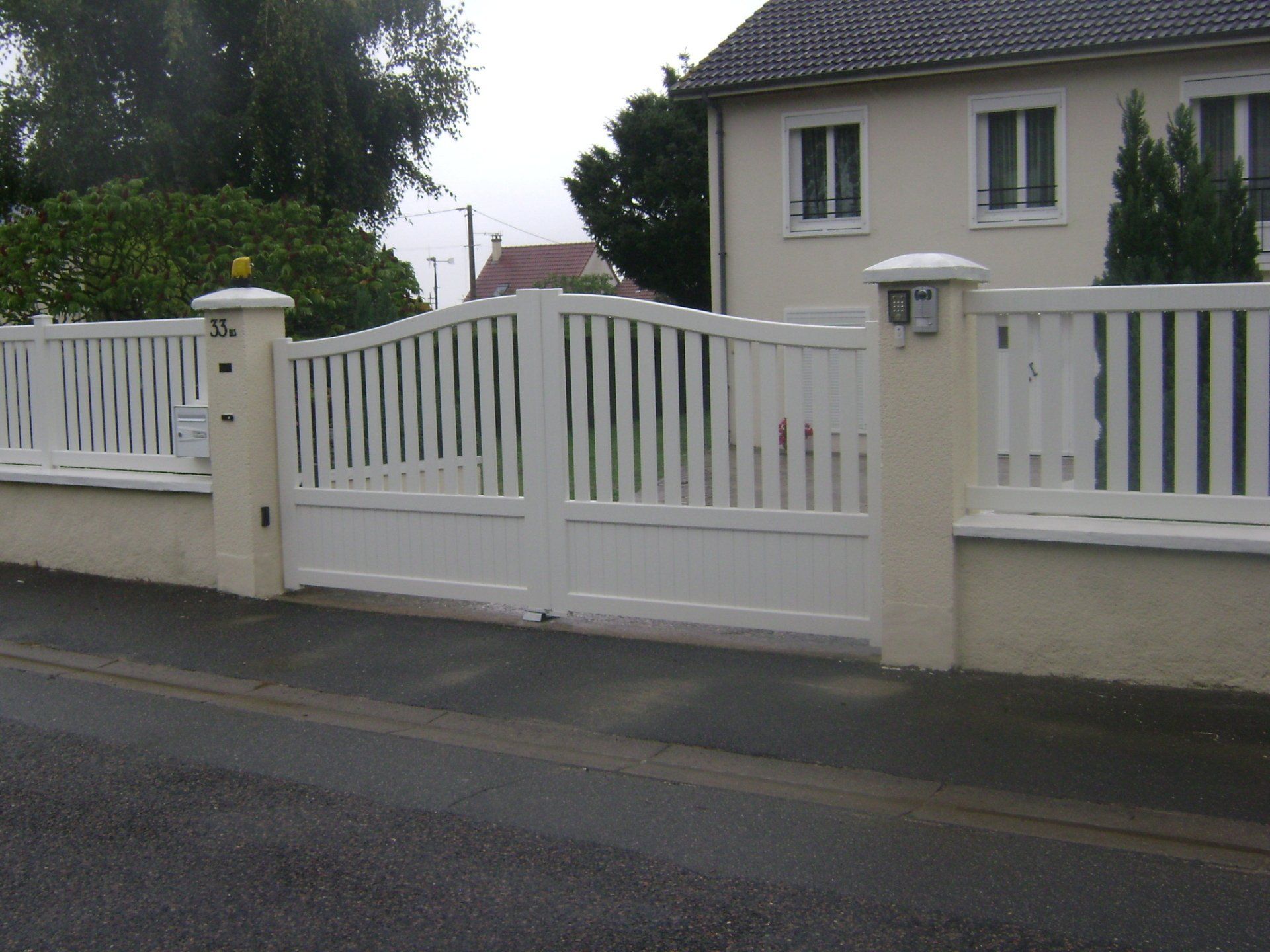 Portail et clôture blancs donnant sur une rue résidentielle, avec une maison beige à l'arrière.