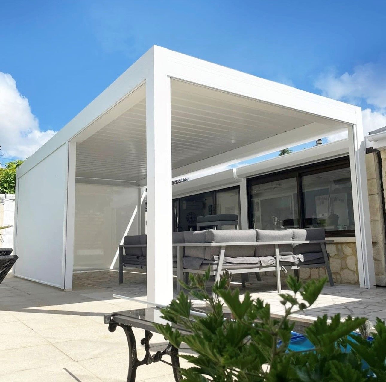Pergola blanche moderne avec terrasse, coin salon extérieur, mur en pierre et ciel bleu éclatant