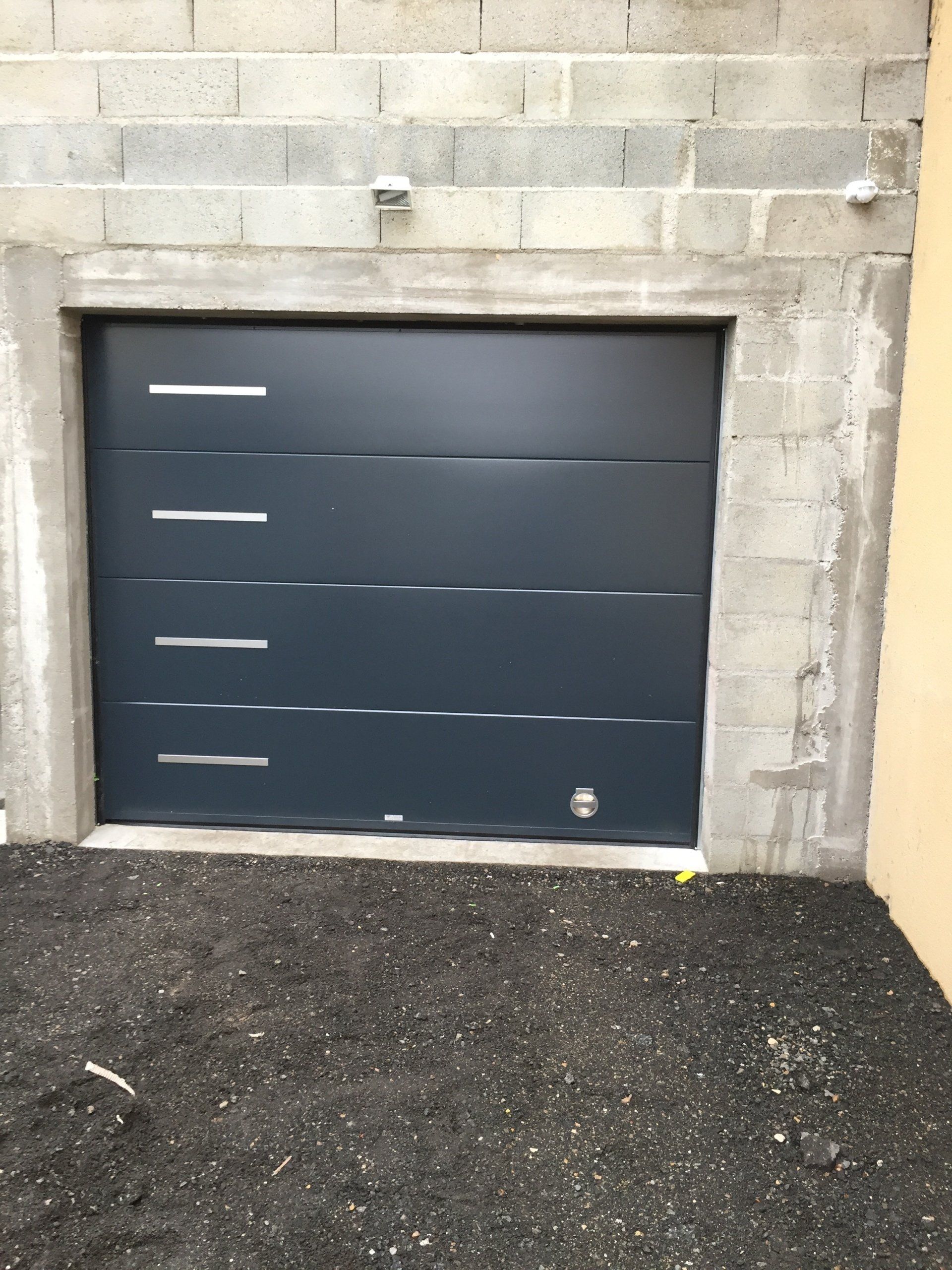 Porte de garage gris foncé encastrée dans un mur en parpaings, avec un sol gravillonné devant.