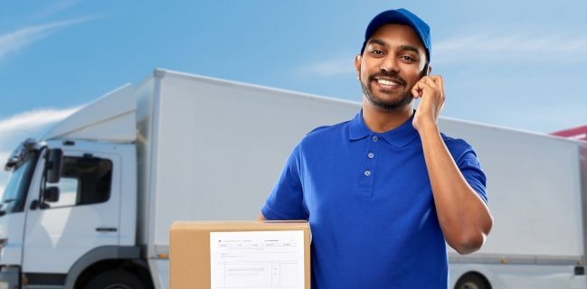 Delivery person in blue uniform smiles while talking on phone, holding a package near a truck.