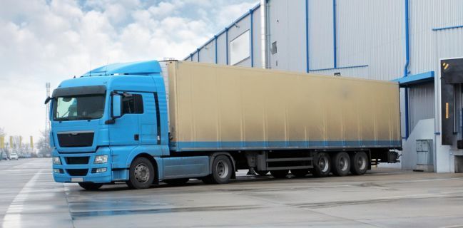 Blue semi-truck parked near a warehouse loading dock on a cloudy day.