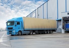 Blue semi-truck parked at a loading dock in front of a warehouse under a cloudy sky.