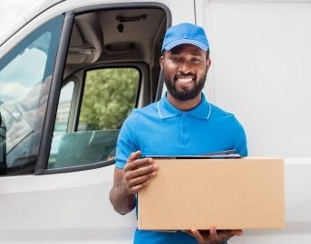 Delivery person in blue uniform holding a cardboard box in front of a white van.