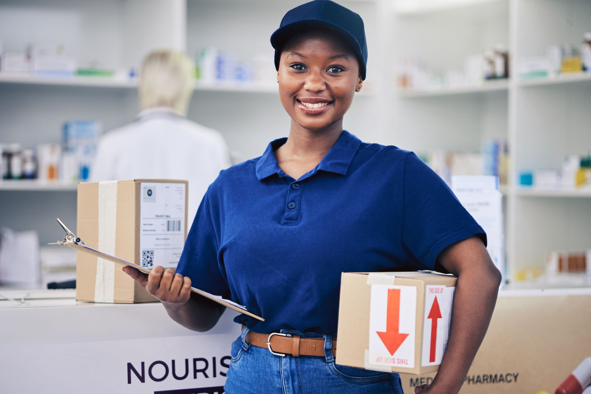 Pharmacy employee holding packages and clipboard, smiling in a pharmacy setting.