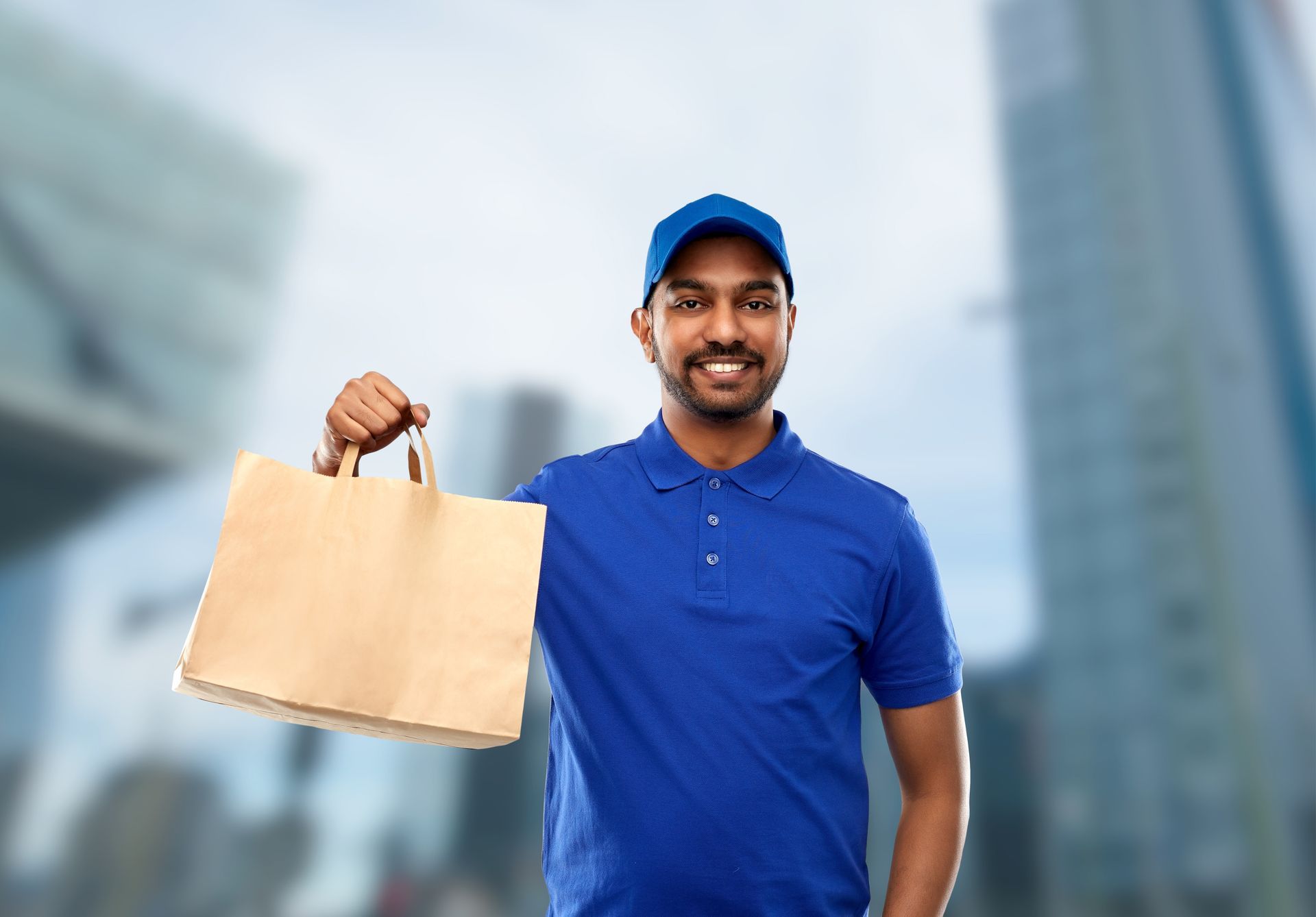 Delivery person holding a brown paper bag, smiling; in front of a city background.