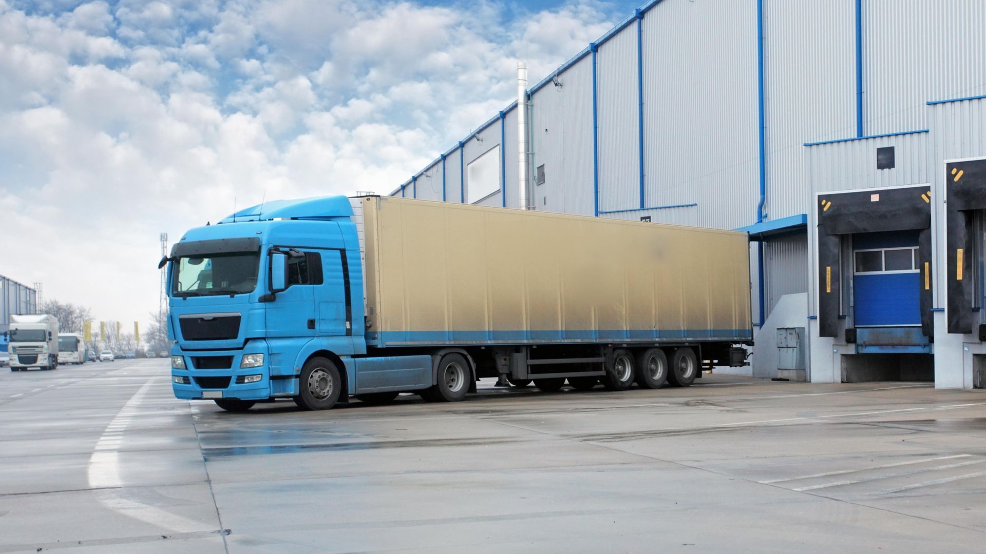 Blue semi-truck backed up to a loading dock at a warehouse under a cloudy sky.