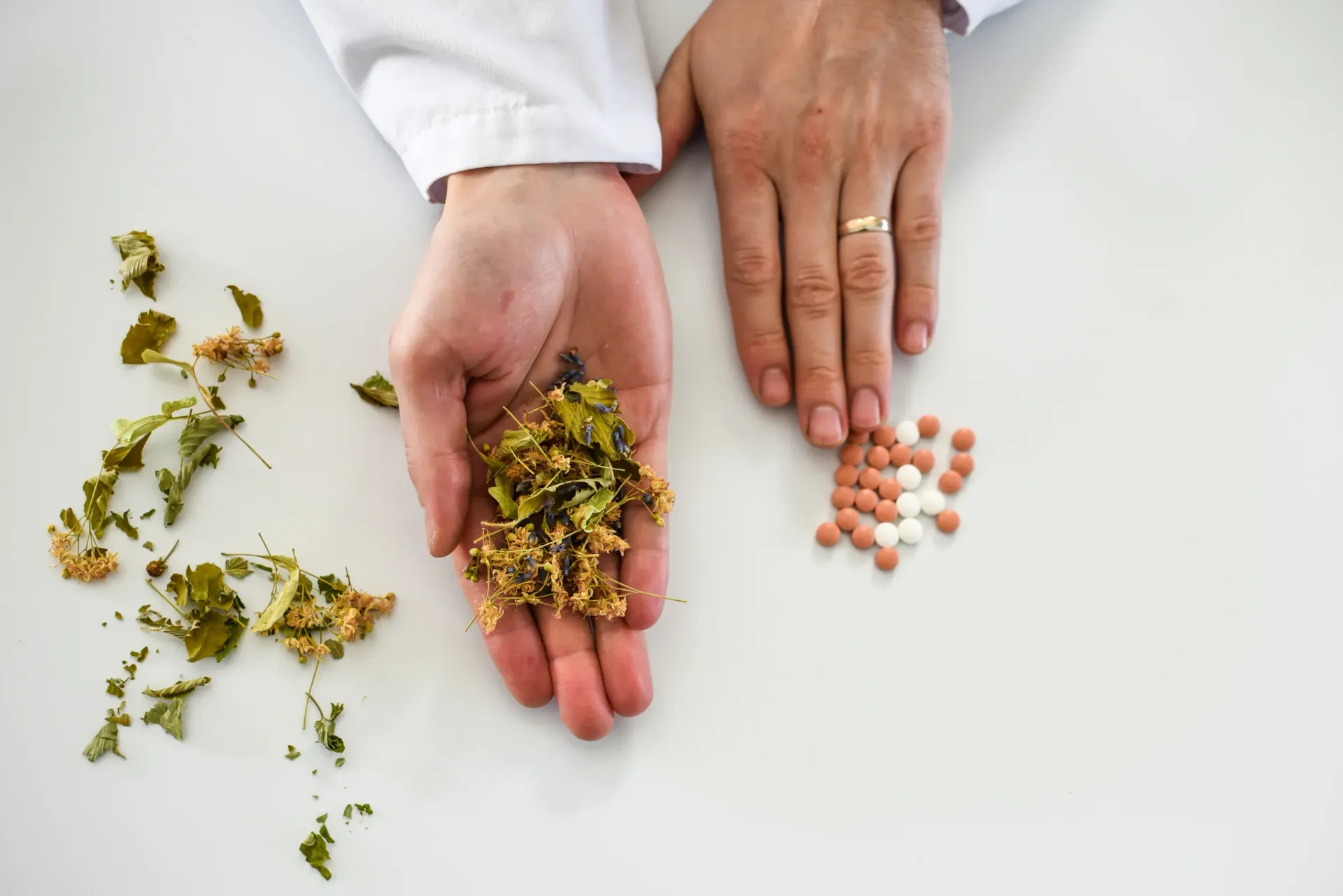 Manos sosteniendo hierbas secas y pastillas sobre una superficie blanca.