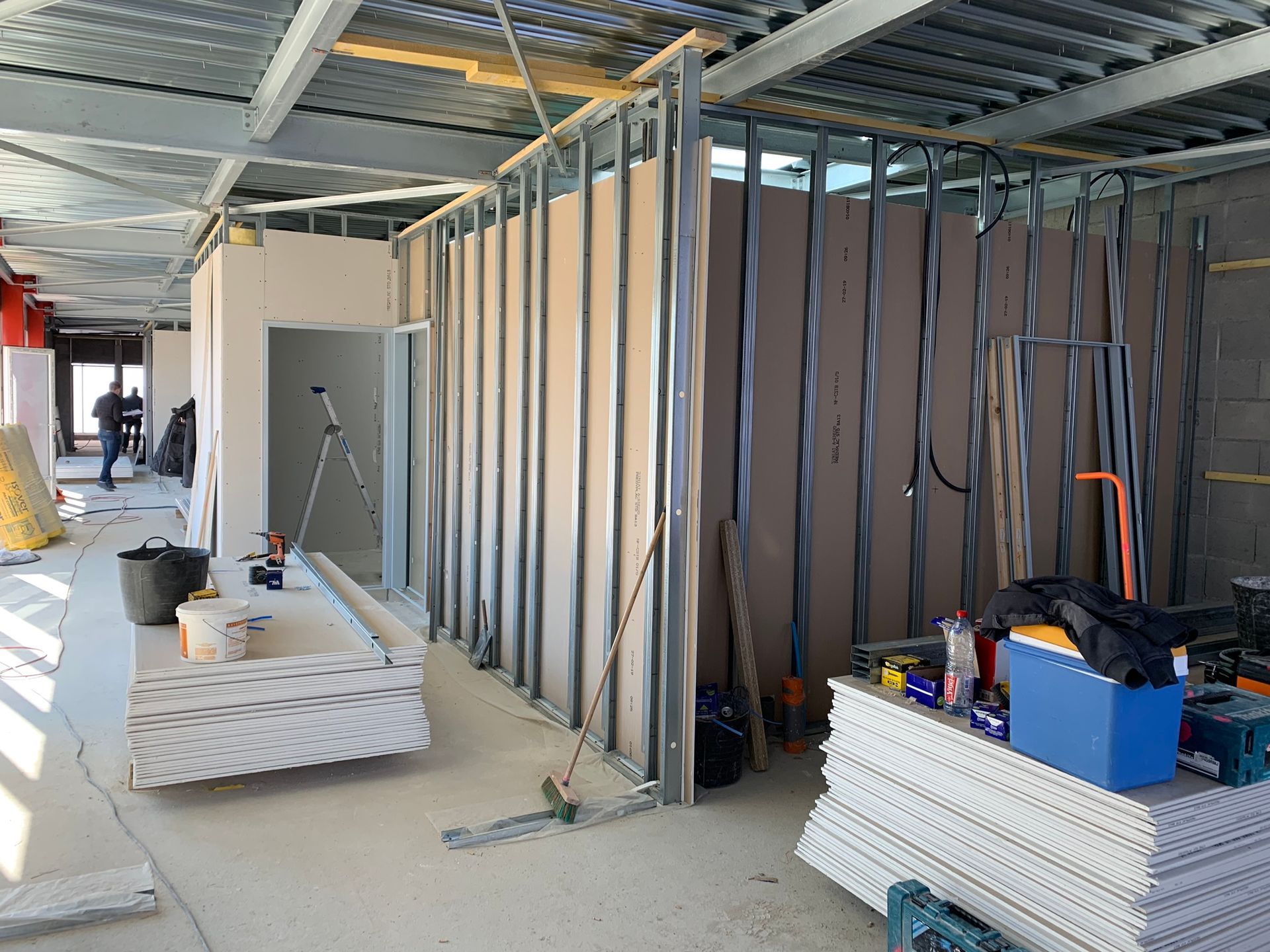 Chantier de construction intérieur avec des murs à ossature métallique, des plaques de plâtre et des outils éparpillés.