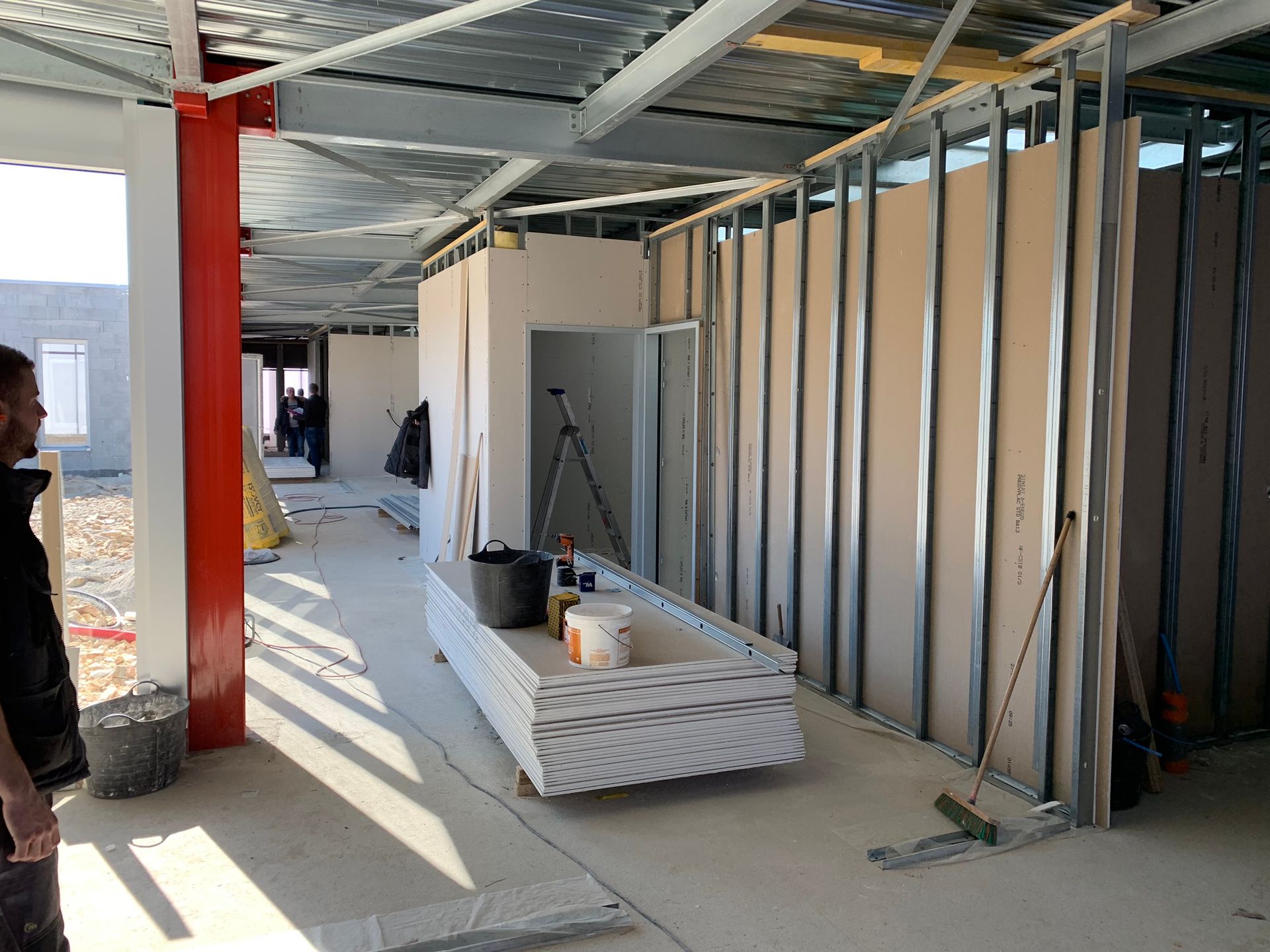 Intérieur d'un chantier ; pose de plaques de plâtre en cours. Montants métalliques, murs non finis, ouvrier visible.