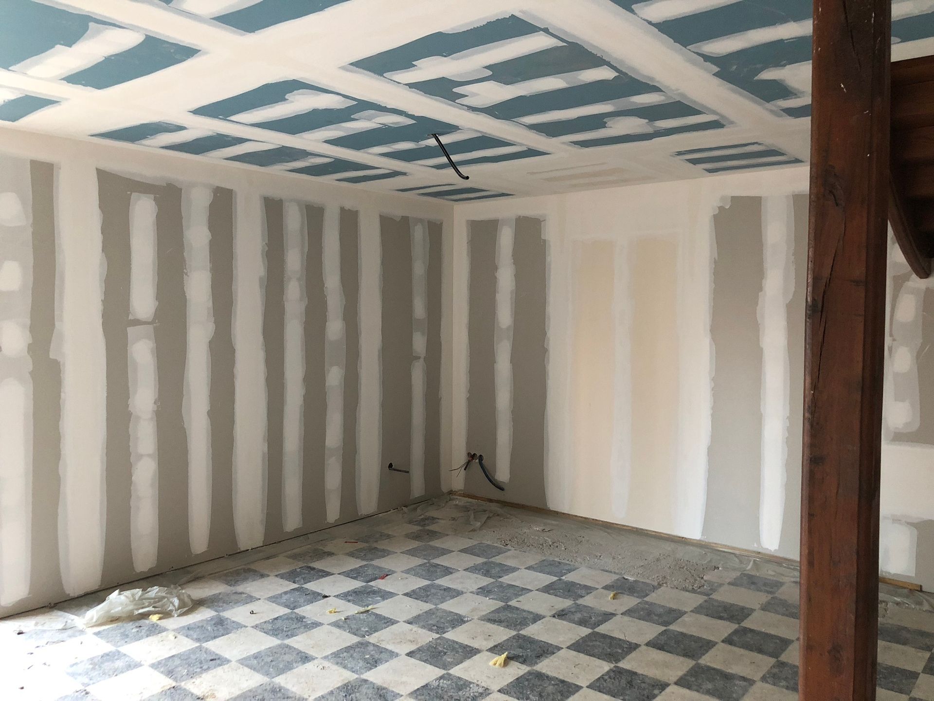 Pièce en travaux avec cloisons sèches apparentes et plafond à caissons bleus. Sol à damier et poutre en bois visibles.