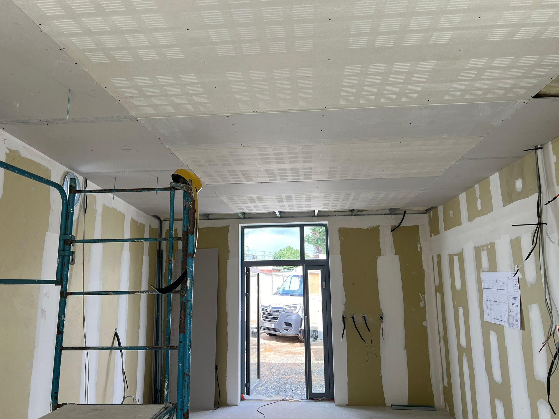 Travaux intérieurs : pièce avec cloisons sèches non finies, échafaudage, porte ouverte et plafond partiellement achevé.
