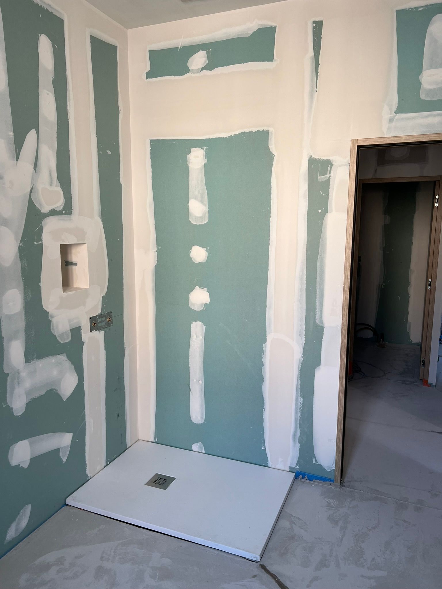 Salle de bains en construction avec receveur de douche et cloisons sèches. Murs bleu-vert, receveur blanc, porte à droite.