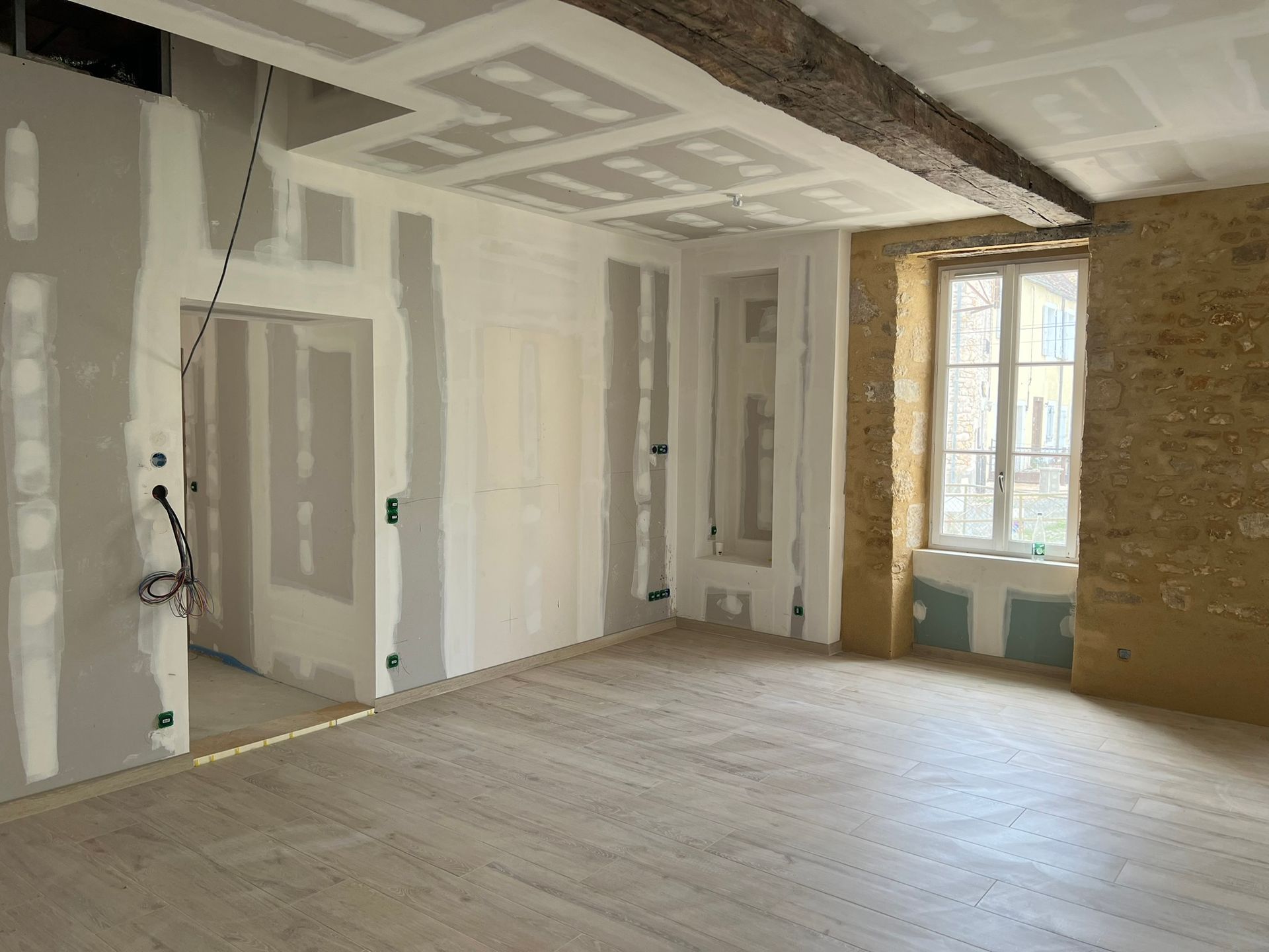 Intérieur d'une pièce en rénovation : cloisons sèches, poutre apparente, parquet et mur en pierre avec une fenêtre.