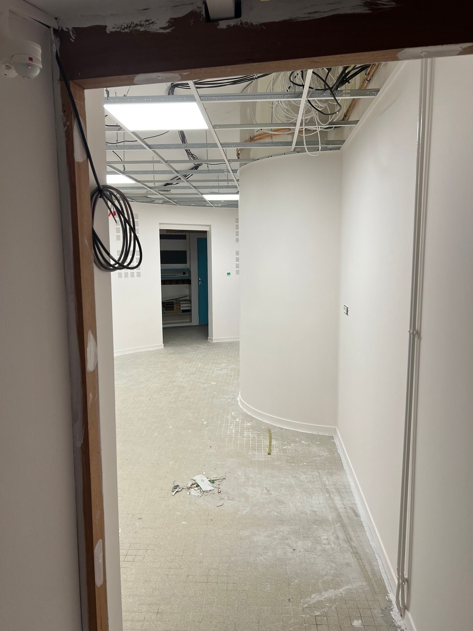 Couloir intérieur en travaux ; murs blancs incurvés, plafond à ossature apparente, débris au sol.