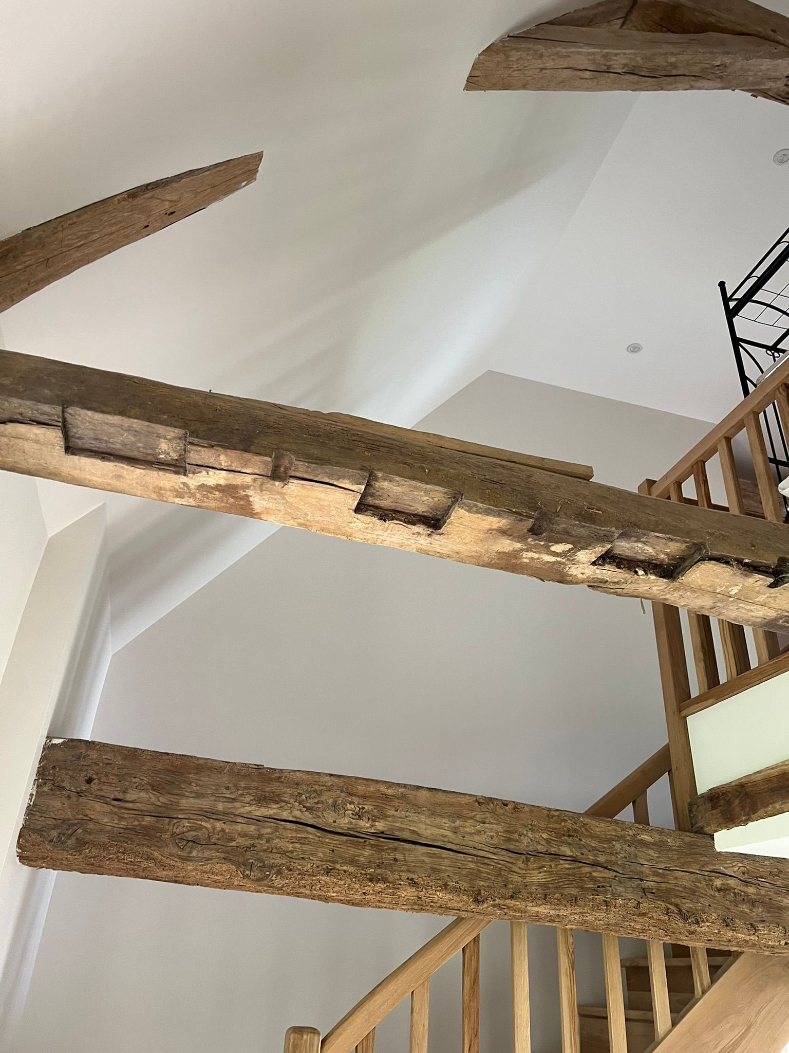 Poutres en bois apparentes dans une pièce aux murs blancs, avec un escalier visible sur la droite.