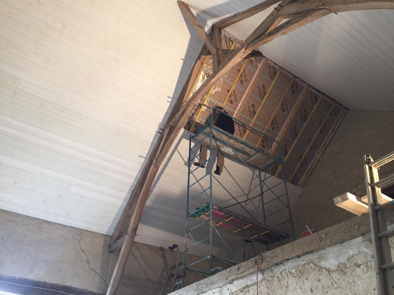 Une personne travaille sur un échafaudage à l'intérieur d'un bâtiment. On aperçoit des poutres en bois et un mur inachevé.