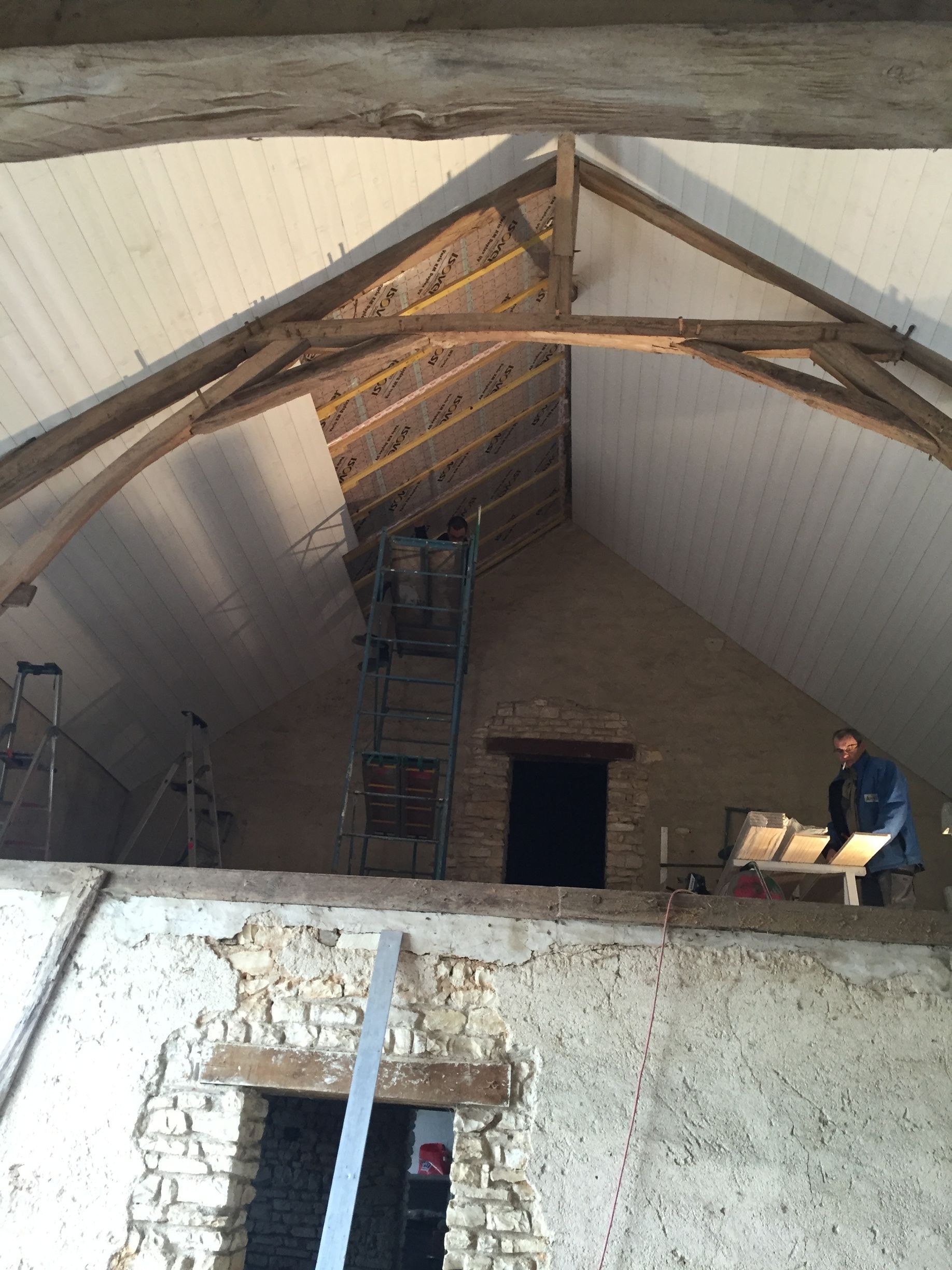 Vue intérieure d'une grange en rénovation. Poutres en bois, plafond partiel et une personne travaillant sur une plateforme.
