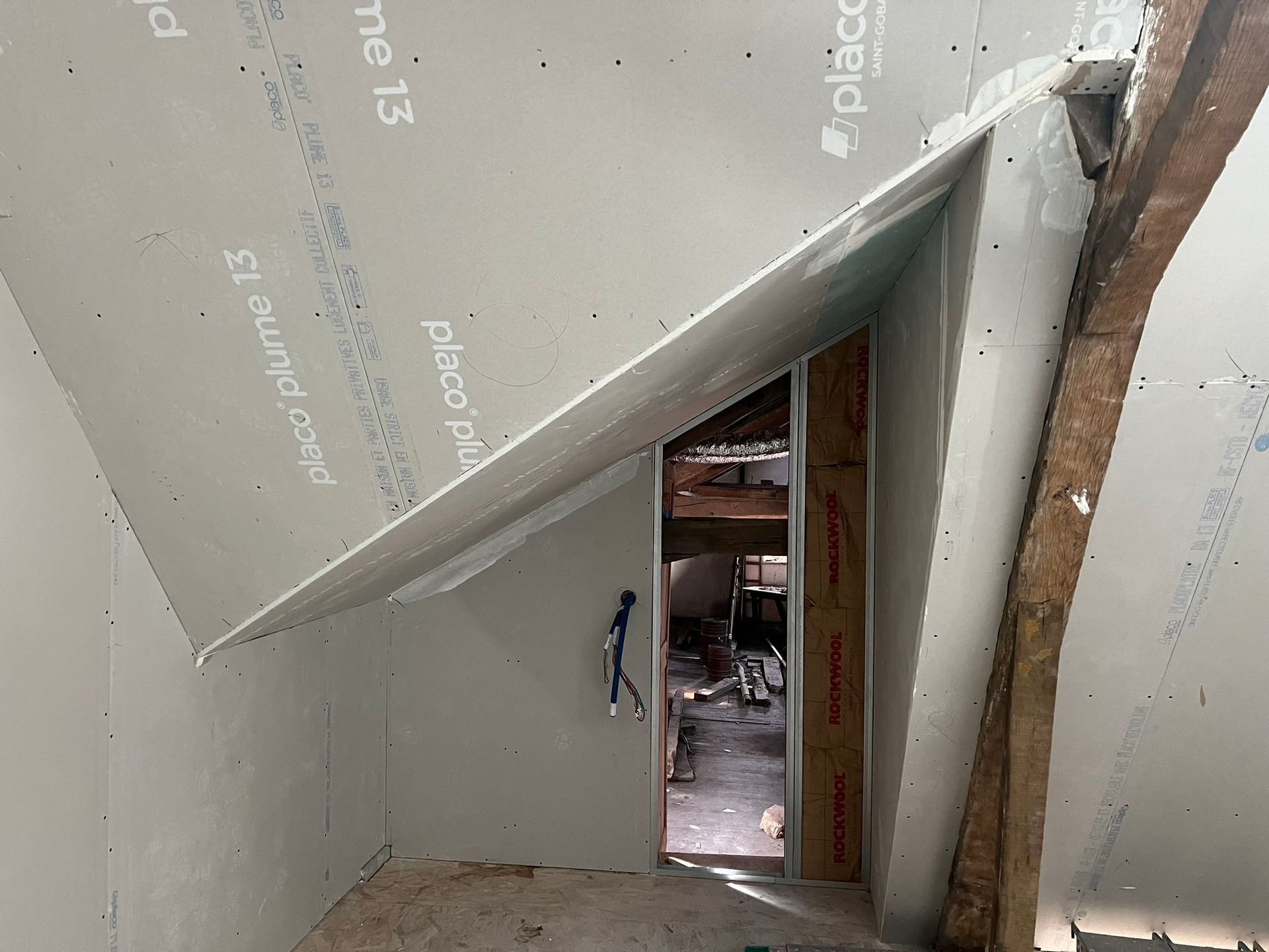 Vue intérieure d'une pièce en construction, avec des poutres en bois apparentes, des cloisons sèches et une porte ouverte.