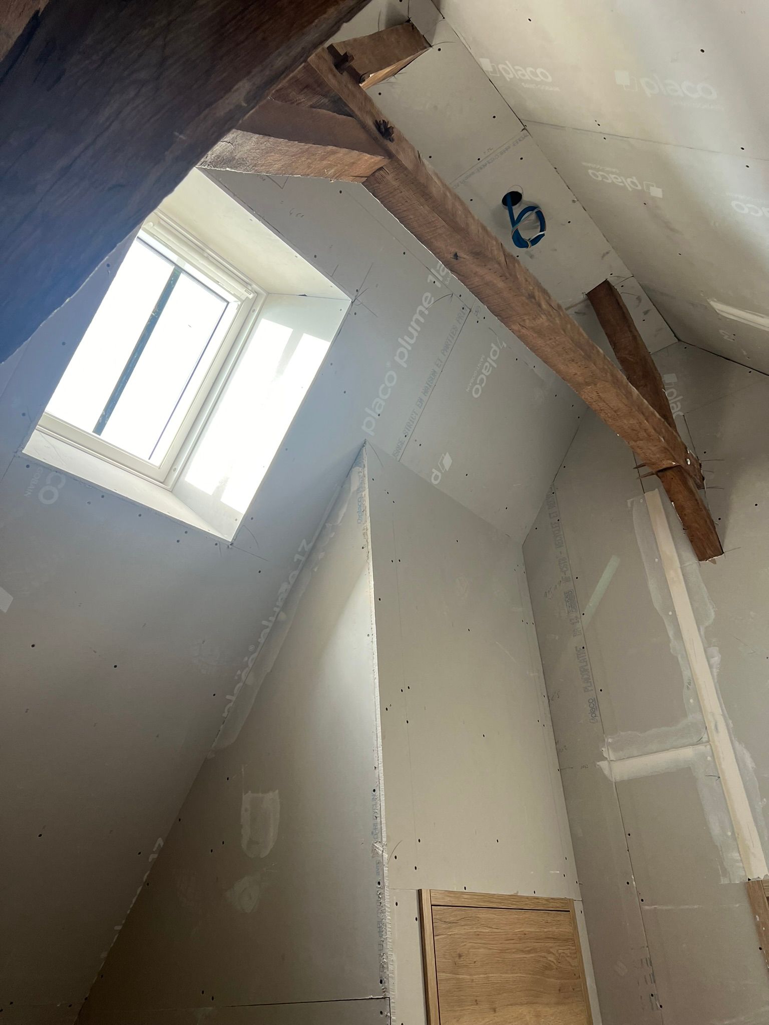 Intérieur du grenier avec poutres apparentes, cloisons sèches et une fenêtre.