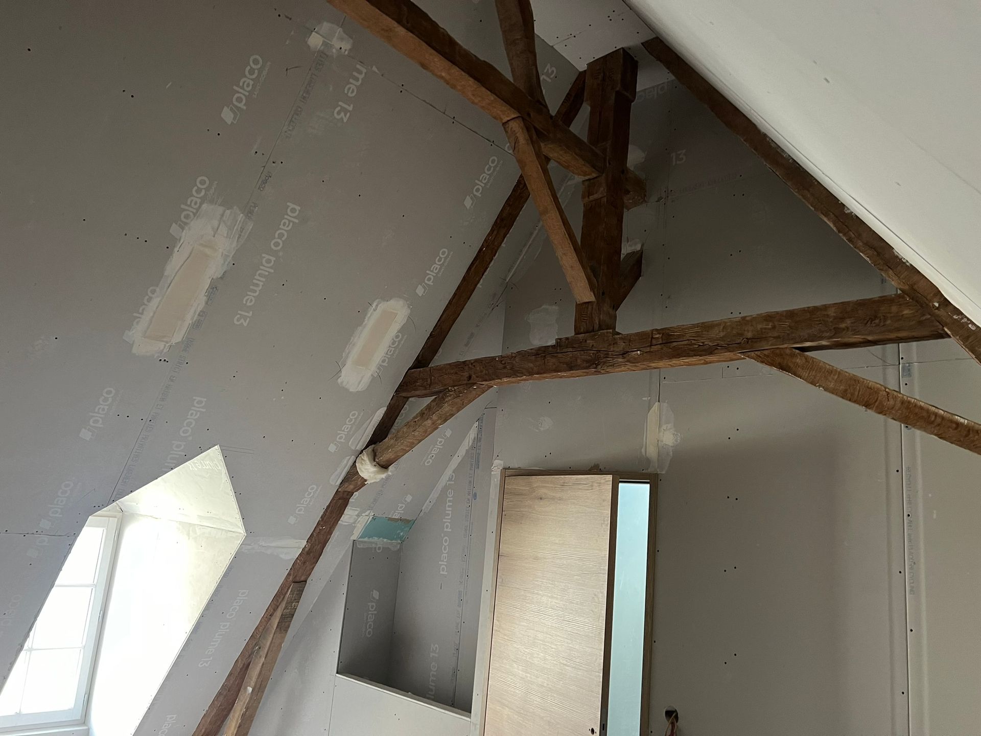 Vue intérieure d'un grenier avec poutres apparentes, cloisons sèches et une petite fenêtre.