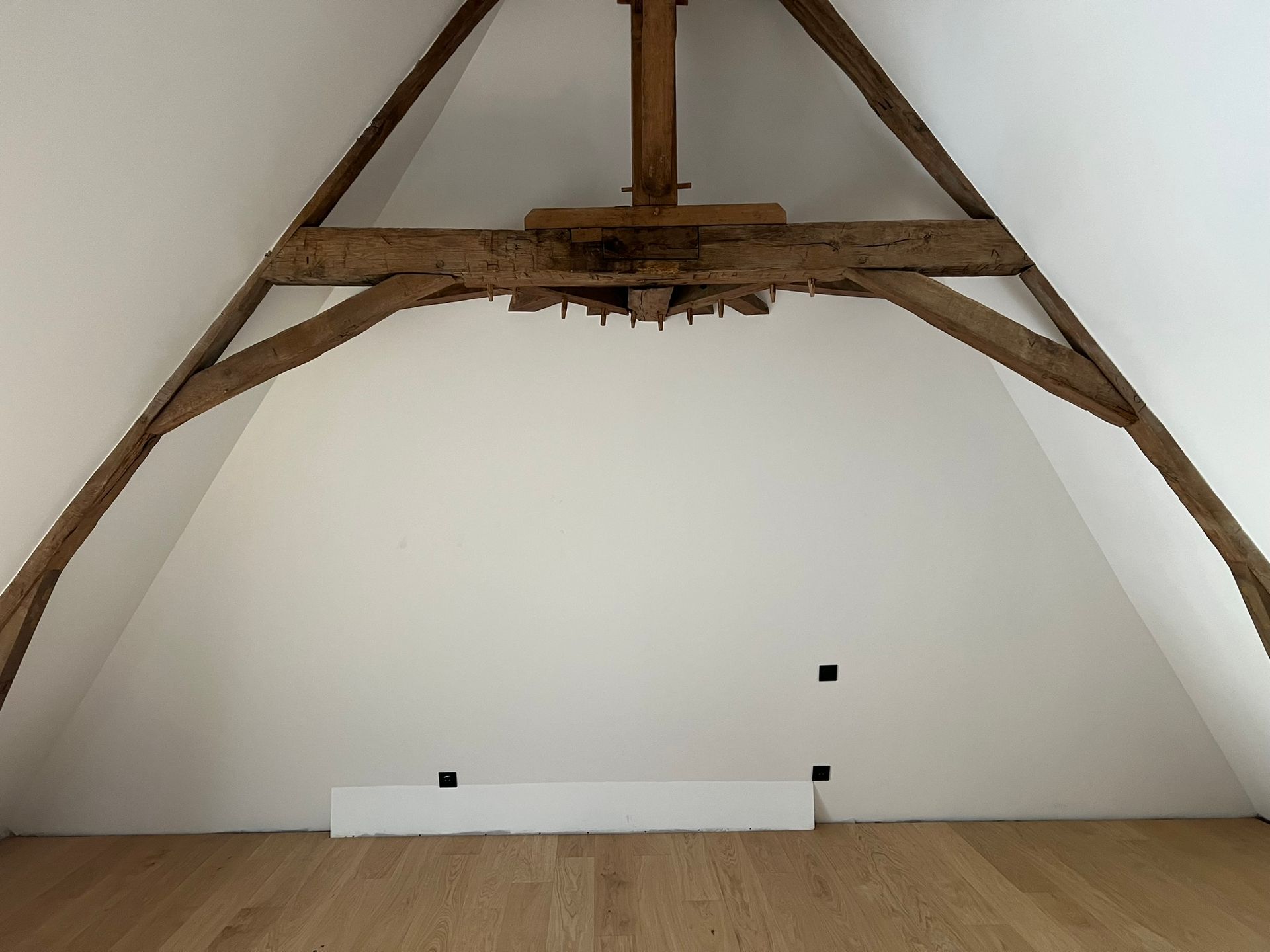 Des poutres en bois soutiennent une pièce aux murs blancs et au parquet clair. Prises électriques murales.