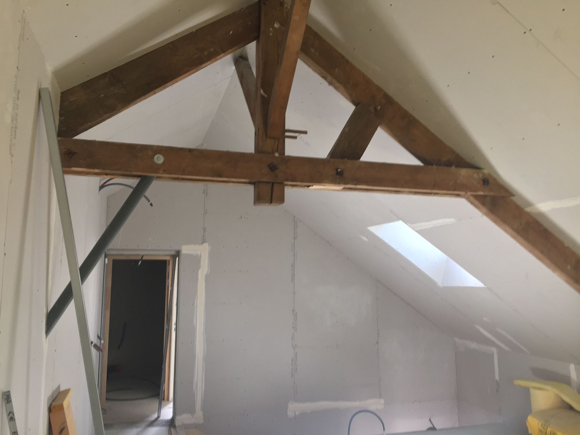 Vue intérieure d'une pièce en construction, avec poutres en bois apparentes, cloisons sèches et puits de lumière.