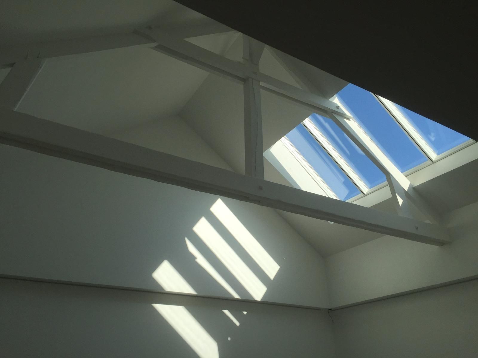 Dans une pièce aux murs blancs, la lumière vive du soleil filtre à travers une lucarne, illuminant l'intérieur.
