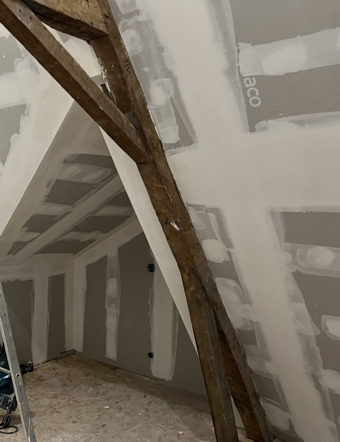 Vue intérieure d'une pièce inachevée avec poutres en bois apparentes, cloisons sèches et un mur partiellement construit.